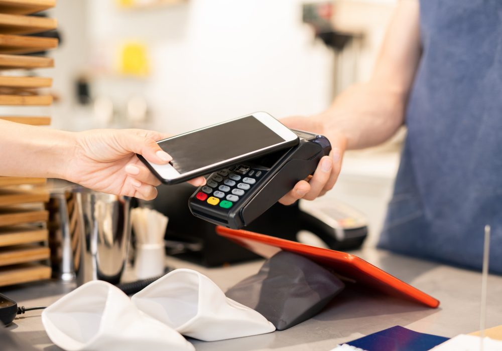 Hand of young woman holding her smartphone close to an electronic payment machine.