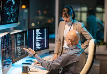 Two intercultural colleagues are having a discussion in front of computer monitors displaying coded data.