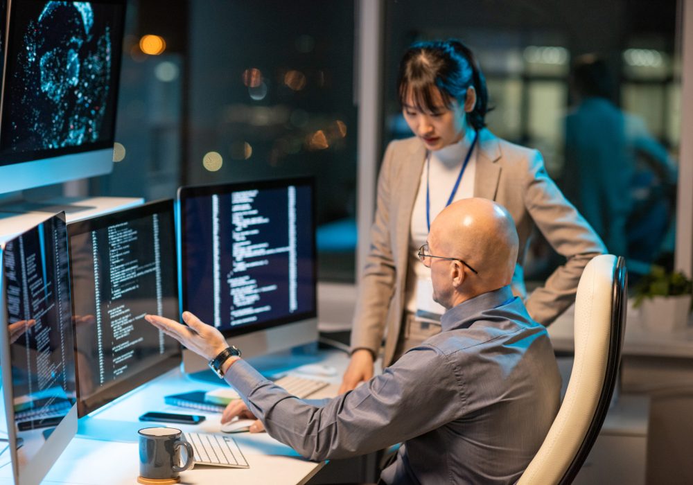 Two intercultural colleagues are having a discussion in front of computer monitors displaying coded data.